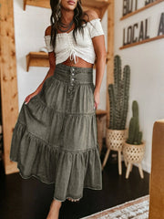 Woman wearing a white off-shoulder top and green tiered skirt in a casual setting with plants.
