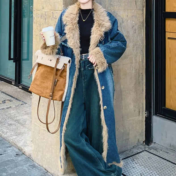 Person wearing a long blue coat with fur trim, holding a brown bag and a white cup, standing against a building.