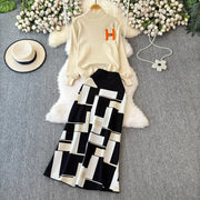 Beige sweater with orange letter 'H' and black and white geometric skirt on a gray surface with flowers and a hat.