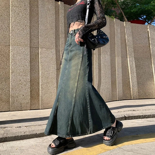 Person wearing a long green denim skirt and black sandals standing on a sidewalk.