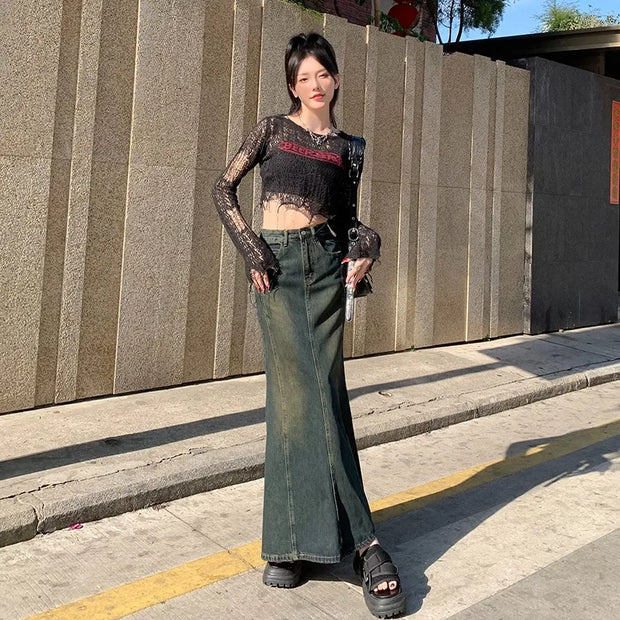 Person wearing a black sheer top with red text and green high-waisted pants standing on a sidewalk.