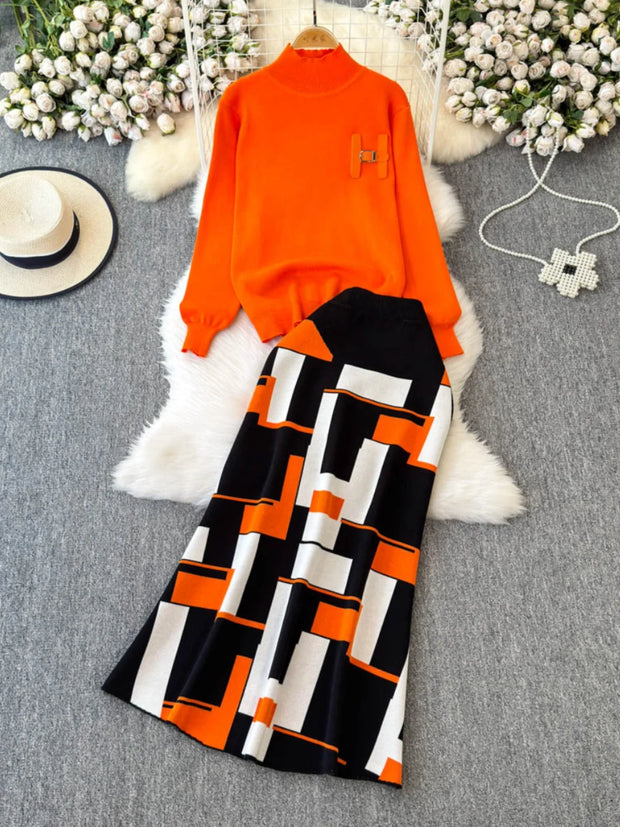 Orange sweater and black and white patterned skirt on a gray surface with flowers and a hat in the background.