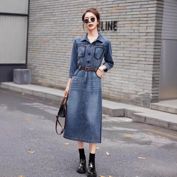 Woman wearing a denim dress standing on a street with a brick wall background.